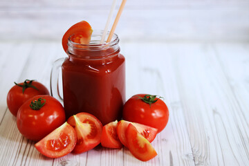 Delicious tomato juice in a glass and fresh tomatoes on a light gray table. Tomato juice on a light background. Fresh juice in a glass with a handle. Glass with juice and a straw. Vegetable drink