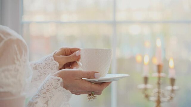 Body part close-up hand holding cup with hot drink tea enjoying taste coffee. woman queen back rear view looking out the window. Fantasy Girl princess Victorian style room, vintage white dress robe