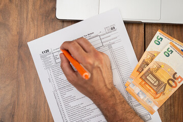 Business owner carefully fills out 1120 tax form on wooden table using laptop and referencing euro bills to ensure all foreign transactions properly recorded