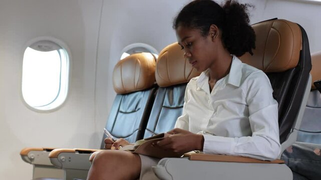 Happy Woman traveling by plane and Write notes while traveling  in airplane. African American business female sitting in airplane during flight in first class seat