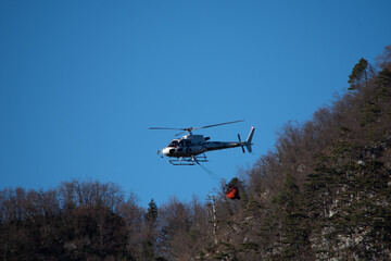 Helicopter in flight for firefighting operation near Illegio (Friuli Venezia Giulia). Water transport with basket, mountains in the background.