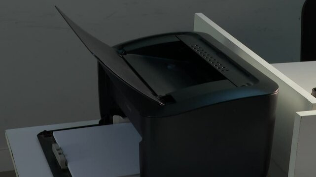 A black office printer sits on a white table, with a blank sheet of paper sticking out. Ready to print important documents for work.