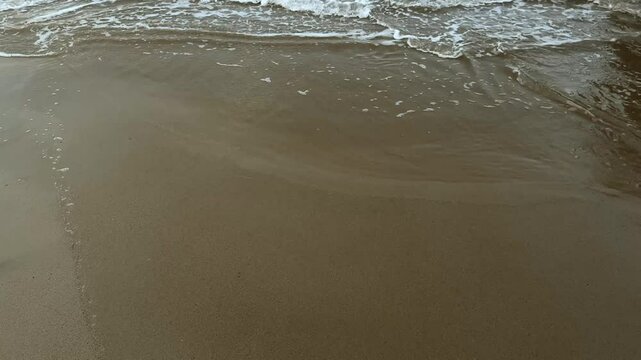 Tranquil sea waves roll onto the beach, forming delicate, abstract foam patterns on wet sand. Peaceful, natural coastal background in soft motion.