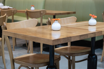 Wooden tables and chairs in a calm cafe setting, adorned with small white vases holding orange flowers, offering a serene space for relaxation or conversation, enhancing ambiance