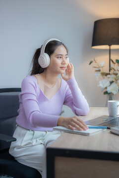 Young Asian businesswoman working from home office and listening to music with headphones while taking notes on a notebook sitting at her desk