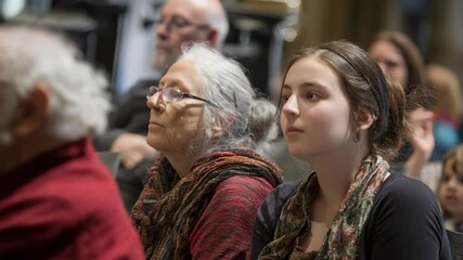 Diverse group of people of varying ages attentively listening, looking upwards in a dimly lit interior space, audience concept.