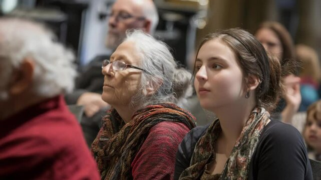Diverse group of people of varying ages attentively listening, looking upwards in a dimly lit interior space, audience concept.