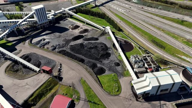 Drone view of an industrial site with coal storage piles, pipelines, sedimentation tanks, and factory buildings near a highway.