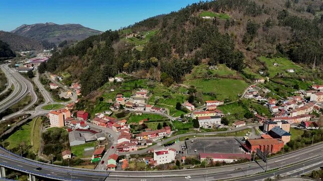 Aerial view of a winding mountain highway passing by a small forest village with scenic hills and a river in the background.