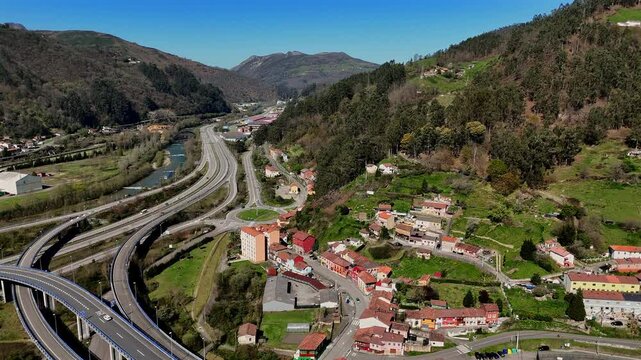 Drone view of a mountain highway interchange running alongside a river and rural village with hills and forest on a clear day.