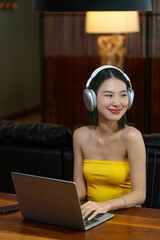 Asian Woman Using Laptop with Headphones at Home. Young Lady Smiling While Working or Streaming...