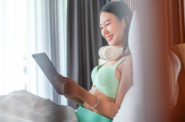 Asian Woman Relaxing in Bed with Tablet and Headphones. Young Female Enjoying Online Entertainment or Learning in Cozy Home Setting.
