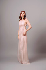 Transformer dress in pastel. Seductive ginger woman in maxi evening gown and high heel shoes on grey studio background. Bridesmaid&rsquo;s summer silk outfit. Gorgeous young sexy lady with curly hairstyle