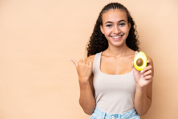 Young woman holding an avocado isolated on beige background pointing to the side to present a product