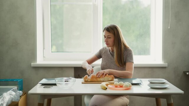 Wide shot of white lady seated on chair carefully slicing cake on plate beside bowl of eggs and stack of plates in modern kitchen with speaker in corner and natural light through window