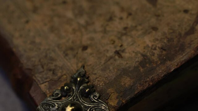 The antique book cover is cracked and dusty, featuring metal ornaments. Slow macro pan. Suitable for historical, educational, or archival projects