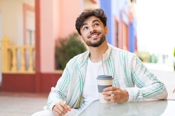 Handsome Arab man holding a take away coffee at outdoors looking up while smiling