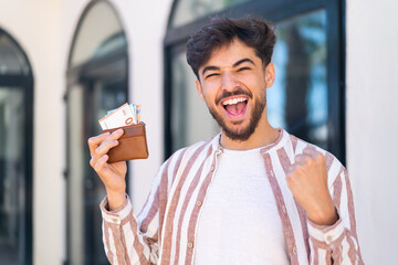 Handsome Arab man at outdoors holding wallet with money and celebrating a victory