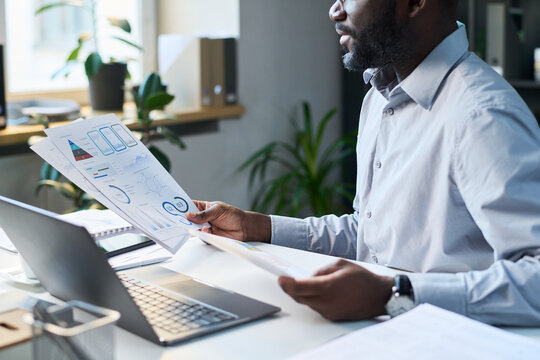 Black male business analyst studying papers with financial graphs and charts while working on report
