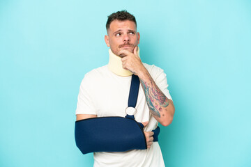 Young caucasian man wearing a sling  and neck lace isolated on blue background having doubts