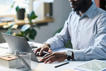 Hands of African American businessman sitting at desk in office, typing on laptop while working on...