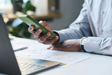 Closeup of hand of unrecognizable black male CEO sitting at desk in office, typing message on smartphone
