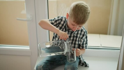 young boy seated barefoot on windowsill wearing checkered shirt and jeans, pouring water from large transparent jar into clear glass cup placed on leg, looking down with focus