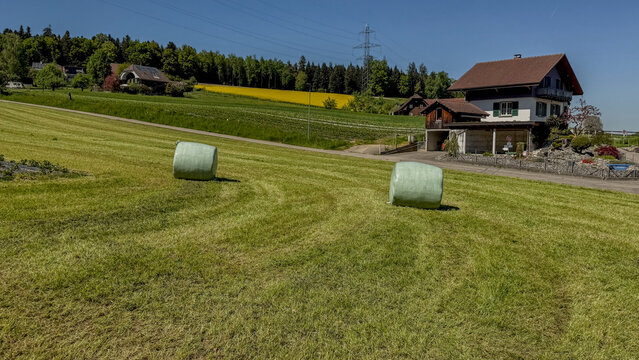Panoramic rural landscape with wrapped hay bales on a freshly cut green field near a traditional farmhouse under a clear blue sky in Switzerland