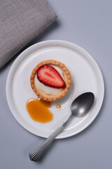Delicate strawberry tart with cream and honey on a white plate. Minimalist dessert style with a spoon on a pastel blue background and a linen napkin. Composition from above.