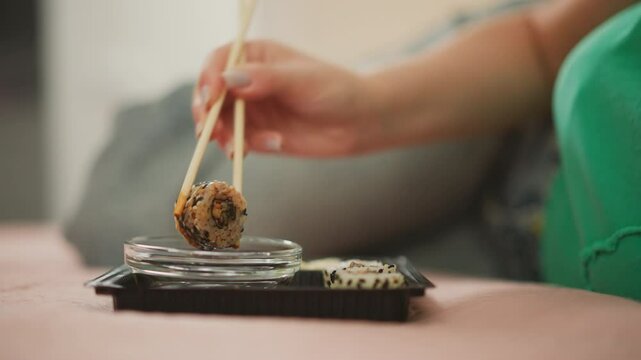 woman with polished nails wearing green shirt lies on bed holding sushi roll with chopsticks dipping it into glass bowl of sauce then lifting it slowly to let sauce drip off with soft blur background