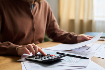 Unrecognizable woman using calculator to check numbers in invoice, while sitting at desk in office