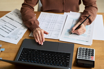 Fototapeta premium High angle of hands of anonymous young woman calculating domestic expenses and utility invoices using laptop, while sitting at desk and managing budget