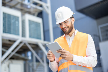 A smiling engineer in a white helmet and safety vest uses a tablet in a industrial setting,...