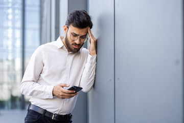 A concerned businessman in glasses looks at his phone while leaning against a building wall...