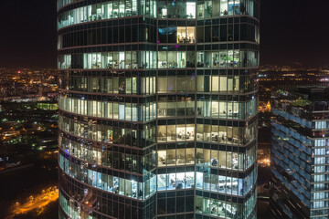 Obraz premium Tall office buildings glow brightly at night, showcasing multiple floors filled with people engaged in work. The vibrant city lights add to the dynamic urban atmosphere.