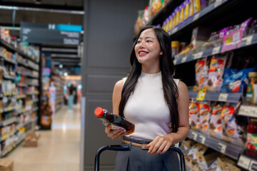 Young woman choosing groceries, holding a bottle and pushing a shopping cart in supermarket