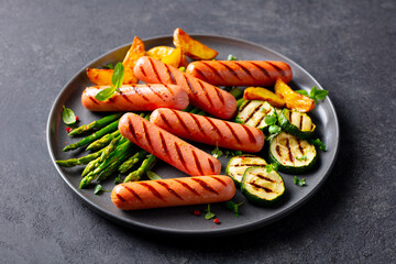 Grilled sausages served with asparagus, zucchini and roasted potatoes, garnished with fresh herbs. Grey background. Close up.