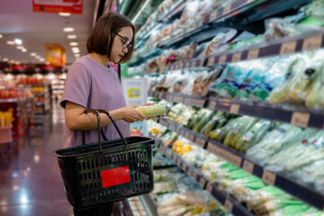 Woman choosing fresh vegetables in supermarket refrigerator shelf, holding shopping basket