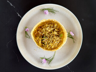 Close up of chicken with torch ginger spicy fried rice with edible flowers, herbs, and local spices. Served with round shape fried wonton skin, and served on a white plate, on a black table.