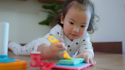 Cute Asian toddler child girl playing with modeling clay on Wooden Floor, enhancing creativity education and motor skills in a cozy home environment.