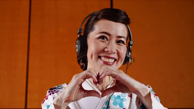 Smiling woman in traditional clothing with headphones making heart gesture with hands indoors