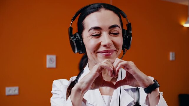 Smiling female doctor wearing headphones and lab coat making heart gesture with hands in clinic setting