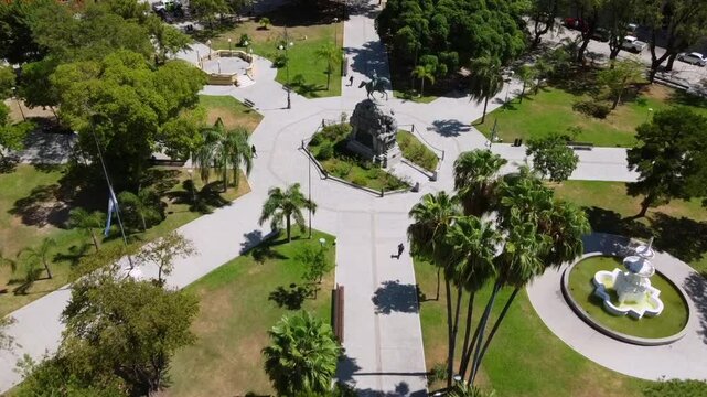 Drone approaches cinematically from above Plaza 25 de Mayo and San Martin statue, flying among palms and vegetation on a sunny day