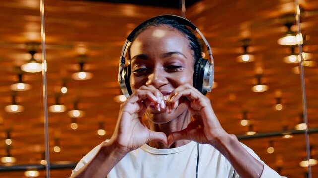 Smiling woman with headphones making heart gesture with hands in stylish modern interior with warm lighting