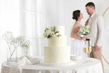 Happy married couple together indoors, focus on wedding cake and glasses of champagne on table