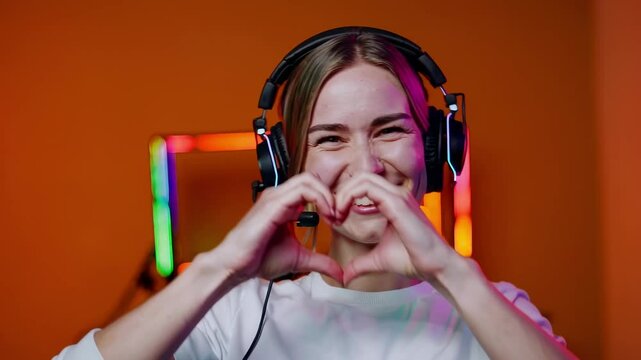 Smiling female gamer with headset making heart gesture with hands in front of colorful RGB-lit gaming setup