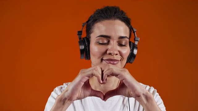 Woman enjoying music with headphones, making heart gesture with hands on vibrant orange background