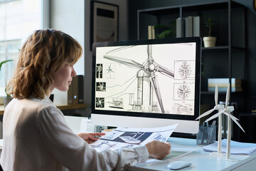 Caucasian female industrial engineer sitting in office in front of computer with technical drawings, working on wind turbine design © pressmaster