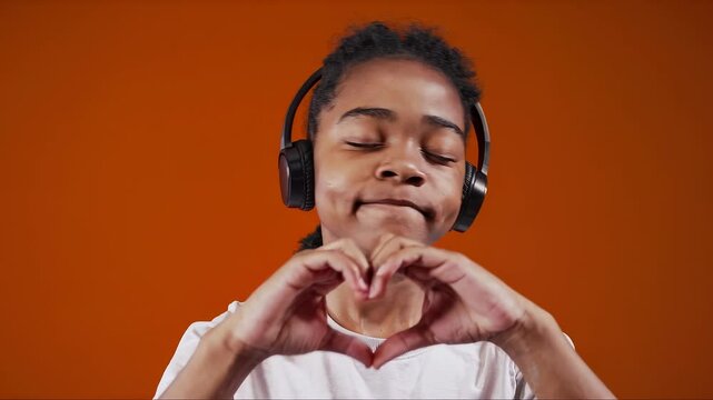 Young boy listening to music with headphones, forming a heart with hands against vibrant orange background