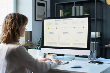 Over shoulder of young Caucasian female manager planning how to lower production costs, using computer in office